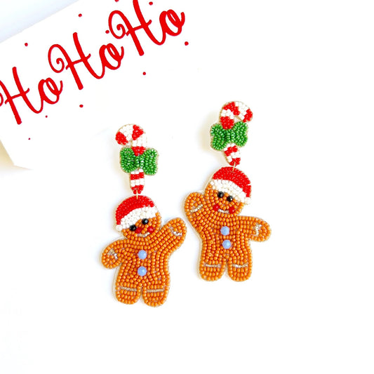 May include: Beaded Christmas gingerbread man earrings.  Each earring features a smiling gingerbread man in shades of orange and brown, wearing a red Santa hat. A candy cane with a green bow is attached to the top of each gingerbread man's head. The earrings are displayed against a white background with the text 'Ho Ho Ho' written in red cursive script.